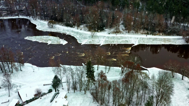 Aerial view of a river flowing through a snow covered landscape with trees during a light snowfall in Latvia. Aerial view of a river flowing through a snow covered landscape with trees during a light snowfall in Latvia.