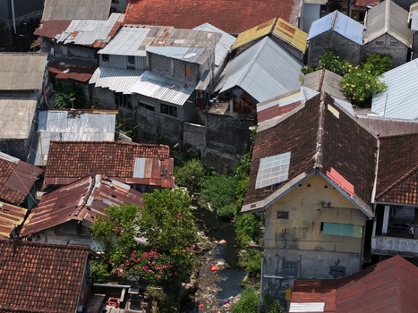 Aerial view of a dense residential neighborhood with tiled and metal rooftops a small stream flowing between houses and lush tropical greenery in Kecamatan Denpasar Barat Bali Indonesia. Aerial view of a dense residential neighborhood with tiled and metal rooftops a small stream flowing between houses and lush tropical greenery in Kecamatan Denpasar Barat Bali Indonesia.