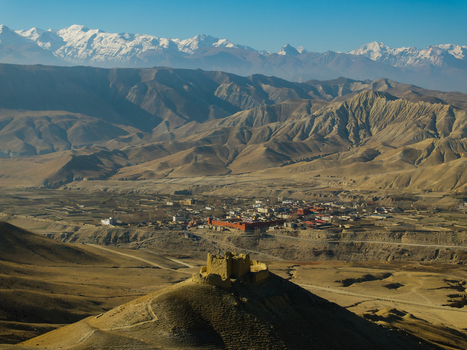 Aerial view of an ancient hill fort overlooking a remote village nestled in the arid landscape of the Himalayas with snow capped peaks in the background in Nepal.