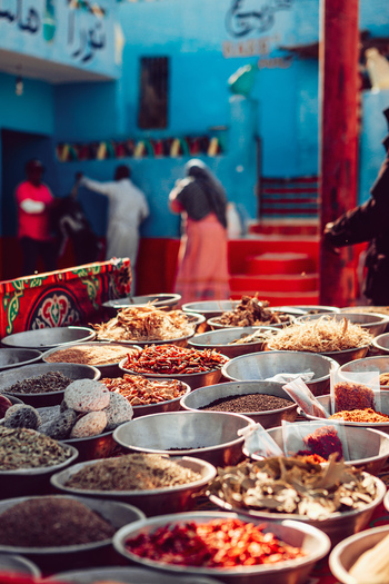 View of various spices and dried herbs displayed in metal bowls at a traditional market with blue buildings and people in the background in Aswan Aswan Governorate Egypt. View of various spices and dried herbs displayed in metal bowls at a traditional market with blue buildings and people in the background in Aswan Aswan Governorate Egypt.