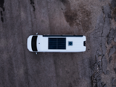 Aerial view of a white camper van with a solar panel on the roof parked on a gravel surface in Latvia.