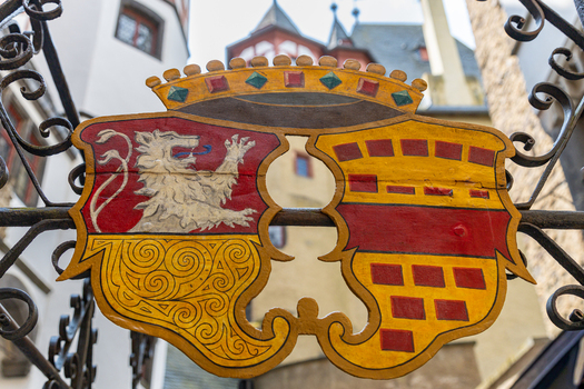 View of the colorful heraldic coat of arms of Eltz Castle mounted on an ornate wrought iron gate with medieval castle towers visible in the blurred background in Wierschem Rhineland Palatinate Germany.