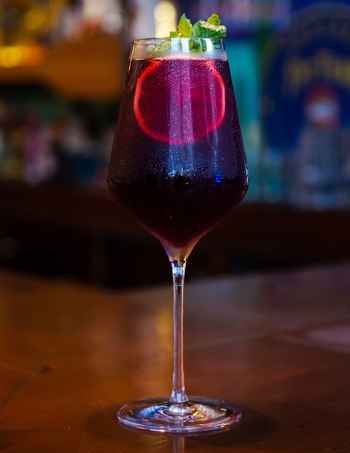 View of a dark red cocktail with a lemon slice and mint leaves in a stemmed glass on a wooden bar counter at Ruba s Bakery Bistro in San Jose del Cabo Baja California Sur Mexico.