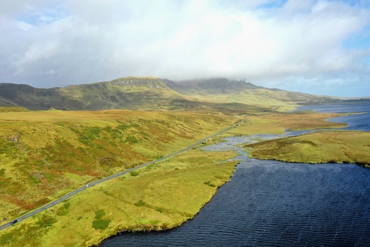 Aerial view of The Storr landscape featuring a winding road along a lake rolling green hills and cloudy skies in Portree Scotland United Kingdom.