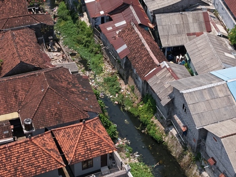 Aerial view of a narrow canal flowing between dense residential buildings with terracotta tiled roofs in Kecamatan Denpasar Barat Bali Indonesia. Aerial view of a narrow canal flowing between dense residential buildings with terracotta tiled roofs in Kecamatan Denpasar Barat Bali Indonesia.