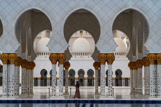 View of the Sheikh Zayed Grand Mosque featuring ornate white arches and floral columns with a person walking in the courtyard in Abu Dhabi Abu Dhabi United Arab Emirates. View of the Sheikh Zayed Grand Mosque featuring ornate white arches and floral columns with a person walking in the courtyard in Abu Dhabi Abu Dhabi United Arab Emirates.