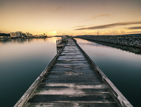 View of the wooden pier extending into the calm waters of Princess Royal Harbour at sunset with industrial structures visible in the distance in Albany WA Australia. View of the wooden pier extending into the calm waters of Princess Royal Harbour at sunset with industrial structures visible in the distance in Albany WA Australia.