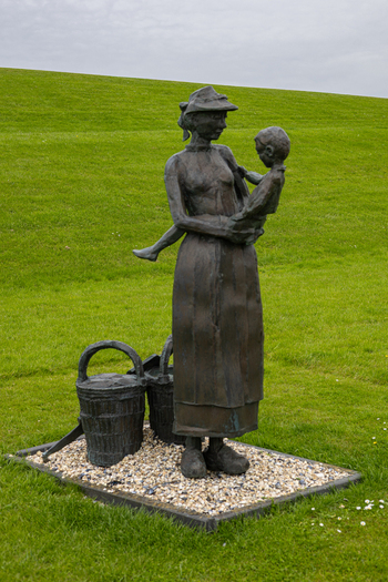View of the bronze De Fiskersfrou statue depicting a woman holding a child with baskets on the ground in Moddergat Friesland Netherlands. View of the bronze De Fiskersfrou statue depicting a woman holding a child with baskets on the ground in Moddergat Friesland Netherlands.