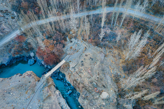 Aerial view of a narrow bridge crossing a turquoise river amidst rugged terrain and skeletal trees Chapursan Valley Hunza Nagar Gilgit Pakistan. Aerial view of a narrow bridge crossing a turquoise river amidst rugged terrain and skeletal trees Chapursan Valley Hunza Nagar Gilgit Pakistan.