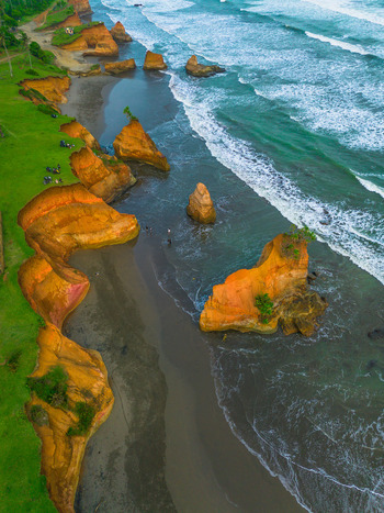 Aerial view of dramatic orange coastal cliffs and rock formations rising from the dark sand beach with turquoise ocean waves crashing against the shore in Bali Indonesia. Aerial view of dramatic orange coastal cliffs and rock formations rising from the dark sand beach with turquoise ocean waves crashing against the shore in Bali Indonesia.