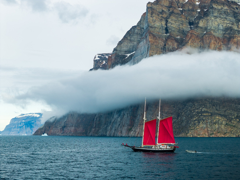 Aerial view of a sailboat with red sails navigating near the towering rocky cliffs of Storoen shrouded in low hanging clouds over the deep blue waters of Avannaata Municipalit Greenland. Aerial view of a sailboat with red sails navigating near the towering rocky cliffs of Storoen shrouded in low hanging clouds over the deep blue waters of Avannaata Municipalit Greenland.