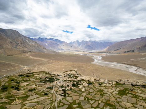 Aerial view of the Zanskar valley featuring patchwork agricultural fields a winding river and dramatic mountain ranges under a cloudy sky in Zanskar Ladakh India.