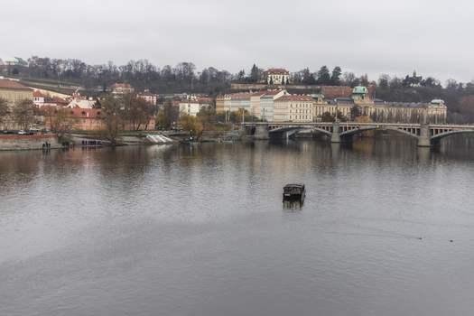 View of the Vltava River with a small boat cruising near the Manes Bridge and historic architecture along the riverbank under a cloudy sky in Prague Prague Czechia. View of the Vltava River with a small boat cruising near the Manes Bridge and historic architecture along the riverbank under a cloudy sky in Prague Prague Czechia.