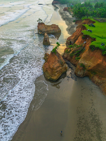 Aerial view of sea stacks and cliffs along the coastline with green grass crashing waves and a person walking on the sandy beach in Bali Indonesia. Aerial view of sea stacks and cliffs along the coastline with green grass crashing waves and a person walking on the sandy beach in Bali Indonesia.