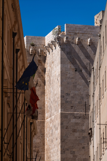 View of the historic Torre dell Elefante stone tower framed by narrow city buildings with flags waving under a clear blue sky in Cagliari Sardegna Italy. View of the historic Torre dell Elefante stone tower framed by narrow city buildings with flags waving under a clear blue sky in Cagliari Sardegna Italy.