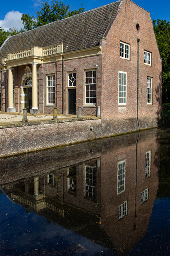 View of Castle Groeneveld with its brick facade and white columns reflected in the calm water of the moat under a blue sky in Baarn Utrecht Netherlands.