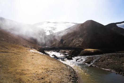 View of a bridge over a shimmering river winding through a rugged landscape with steaming vents rising against the backdrop of stark mountains Hrunamannahreppur Iceland.