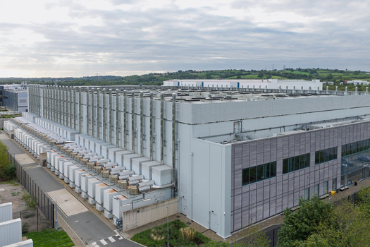 Aerial view of a large data center facility with rows of cooling units on the roof and surrounding green fields in the United Kingdom.