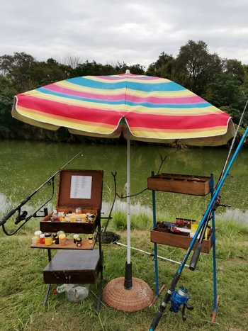 View of fishing gear rods and bait containers under a colorful striped umbrella on the grassy bank of the Crocodile River in Hartbeespoort North West South Africa. View of fishing gear rods and bait containers under a colorful striped umbrella on the grassy bank of the Crocodile River in Hartbeespoort North West South Africa.