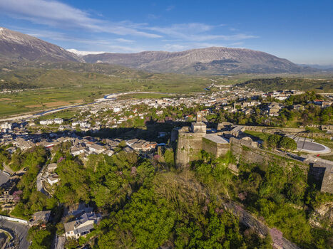 Aerial view of Gjirokaster Castle overlooking the historic stone city with lush green hills and distant mountains under a clear sky in Gjirokaster Gjirokaster County Albania. Aerial view of Gjirokaster Castle overlooking the historic stone city with lush green hills and distant mountains under a clear sky in Gjirokaster Gjirokaster County Albania.