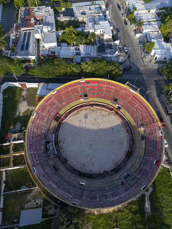 Aerial view of Plaza de Toros Merida showing the circular arena with red and yellow seating tiers surrounded by urban streets and lush trees Merida Yucatan Mexico. Aerial view of Plaza de Toros Merida showing the circular arena with red and yellow seating tiers surrounded by urban streets and lush trees Merida Yucatan Mexico.
