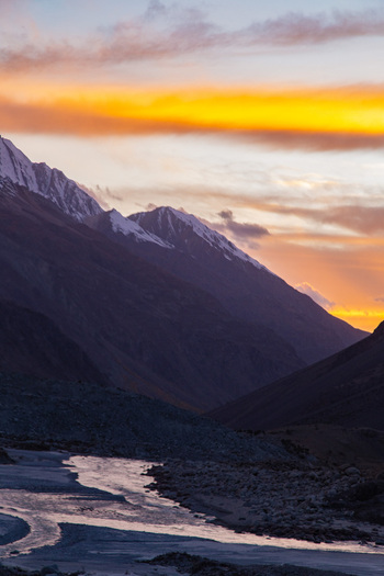 View of a dramatic mountain landscape at twilight where a winding icy river reflects the fiery orange and yellow sky contrasting with the dark snow capped peaks Skardu Pakistan. View of a dramatic mountain landscape at twilight where a winding icy river reflects the fiery orange and yellow sky contrasting with the dark snow capped peaks Skardu Pakistan.