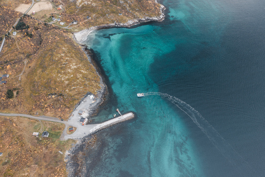 Aerial view of the Senja coastline with a small boat leaving a stone pier surrounded by turquoise waters and rugged terrain in Senja Troms Norway.