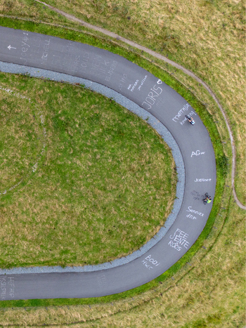 Aerial view of Col du Vam with cyclists riding on the curved asphalt road marked with graffiti and names surrounded by green grassy hills in Wijster Drenthe Netherlands. Aerial view of Col du Vam with cyclists riding on the curved asphalt road marked with graffiti and names surrounded by green grassy hills in Wijster Drenthe Netherlands.