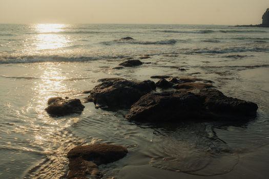 View of the sun kissing the ocean horizon as waves lap against dark rocks on a sandy shore casting golden reflections Chiplun Maharashtra India. View of the sun kissing the ocean horizon as waves lap against dark rocks on a sandy shore casting golden reflections Chiplun Maharashtra India.
