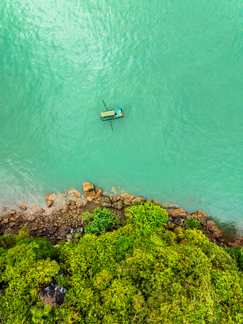 Aerial view of a small boat floating on turquoise water near a rocky forested coastline in Vietnam. Aerial view of a small boat floating on turquoise water near a rocky forested coastline in Vietnam.