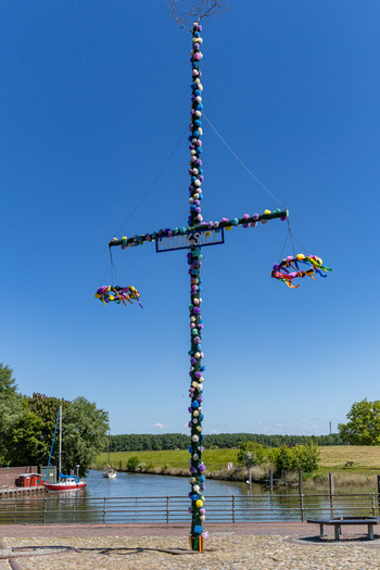View of the decorated Hooksiel maypole with colorful wreaths and flowers standing by a canal with boats under a clear blue sky in Wangerland Niedersachsen Germany. View of the decorated Hooksiel maypole with colorful wreaths and flowers standing by a canal with boats under a clear blue sky in Wangerland Niedersachsen Germany.