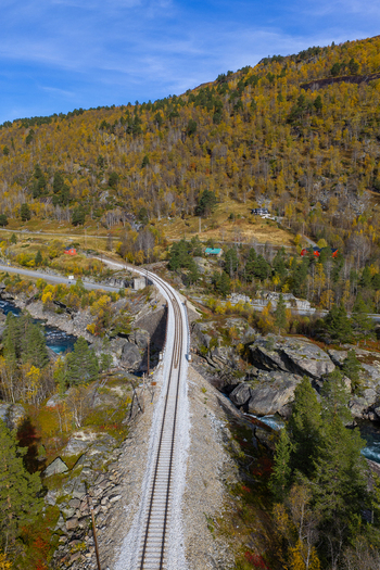 Aerial view of a railway track crossing a bridge over a river surrounded by autumn colored forests and rocky terrain in Bjorli Innlandet Norway. Aerial view of a railway track crossing a bridge over a river surrounded by autumn colored forests and rocky terrain in Bjorli Innlandet Norway.