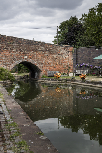 View of a historic brick bridge over a calm canal with reflections in the water stone towpath wooden benches and colorful flowers in Coventry England United Kingdom.