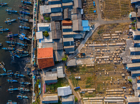 Aerial view of fishing boats docked along a pier next to a coastal village with houses and drying racks in Cilacap Regency Central Java Indonesia. Aerial view of fishing boats docked along a pier next to a coastal village with houses and drying racks in Cilacap Regency Central Java Indonesia.