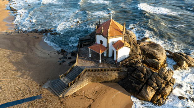 Aerial view of Capela do Senhor da Pedra built on a rocky outcrop with people on the stairs surrounded by crashing waves and a sandy beach in Miramar Porto Portugal.