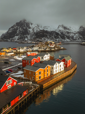 Aerial view of vibrant red and yellow buildings contrasting against the dark reflective waters and snow dusted mountains of Henningsv&aelig;r Nordland Norway.