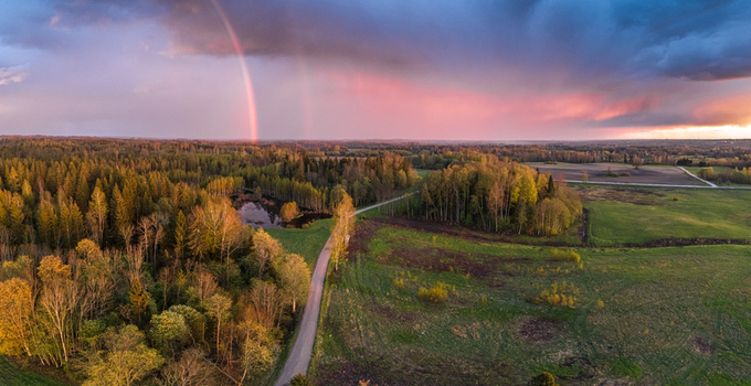 Aerial view of a rural landscape featuring a winding road through lush forests and green fields with a small pond and a vibrant rainbow Latvia. Aerial view of a rural landscape featuring a winding road through lush forests and green fields with a small pond and a vibrant rainbow Latvia.