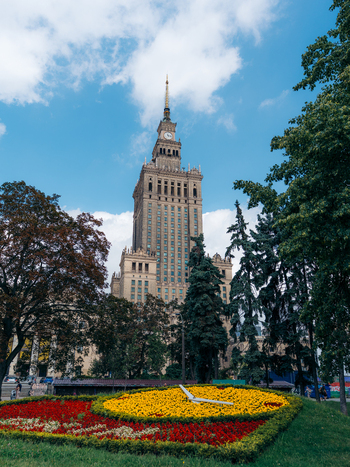 View of the Palace of Culture and Science rising above a vibrant floral clock and lush trees under a bright blue sky in Warszawa Mazowieckie Poland. View of the Palace of Culture and Science rising above a vibrant floral clock and lush trees under a bright blue sky in Warszawa Mazowieckie Poland.