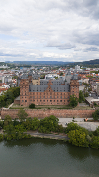 Aerial view of Schloss Johannisburg a historic red sandstone palace on the banks of the Main River surrounded by lush trees and the urban landscape of Aschaffenburg Bayern Germany.