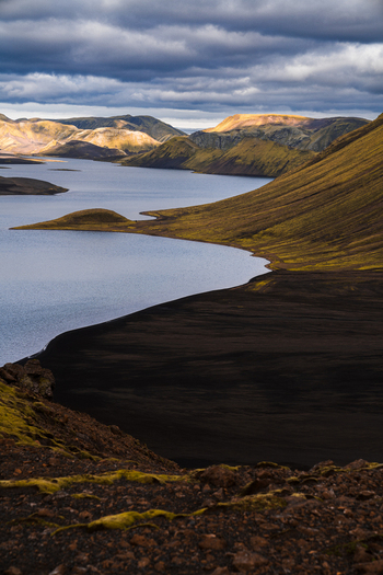 View of the serene lake reflecting cloudy skies nestled amidst rolling hills with dark volcanic soil and vibrant green moss Skaftárhreppur Iceland. View of the serene lake reflecting cloudy skies nestled amidst rolling hills with dark volcanic soil and vibrant green moss Skaftárhreppur Iceland.