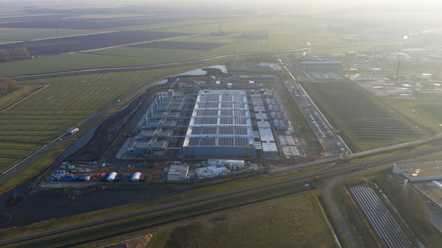 Aerial view of a large industrial data center facility with solar panels on the roof surrounded by green fields and canals under a hazy morning sky in Winschoten Groningen Netherlands.