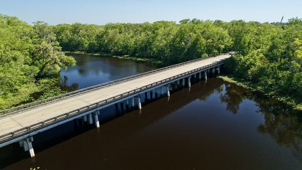 Aerial view of Sixmile Creek bridge crossing dark river water surrounded by dense green forest and lush vegetation in St. Johns County Florida United States.
