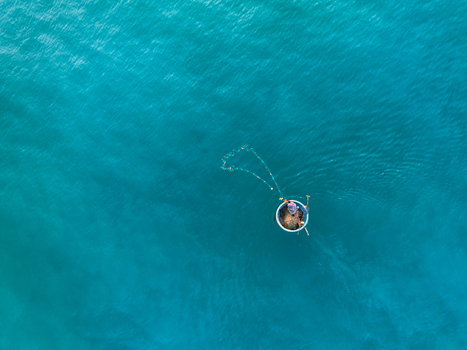 Aerial view of a fisherman casting a net from a traditional round basket boat on the turquoise waters of Vietnam. Aerial view of a fisherman casting a net from a traditional round basket boat on the turquoise waters of Vietnam.