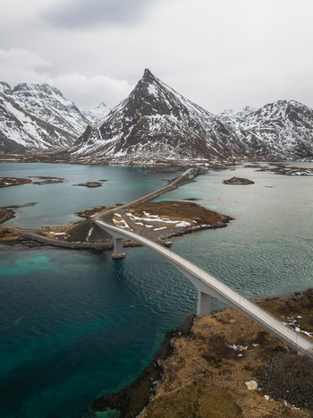 Aerial view of Flakstadbruene bridge connecting islands amidst turquoise waters with snow capped mountains piercing the skyline Flakstadbruene Nordland Norway. Aerial view of Flakstadbruene bridge connecting islands amidst turquoise waters with snow capped mountains piercing the skyline Flakstadbruene Nordland Norway.