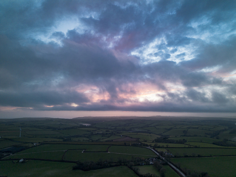 Aerial view of the rural landscape with green fields hedgerows and a wind turbine under a dramatic cloudy sky at dusk in Looe Cornwall United Kingdom. Aerial view of the rural landscape with green fields hedgerows and a wind turbine under a dramatic cloudy sky at dusk in Looe Cornwall United Kingdom.