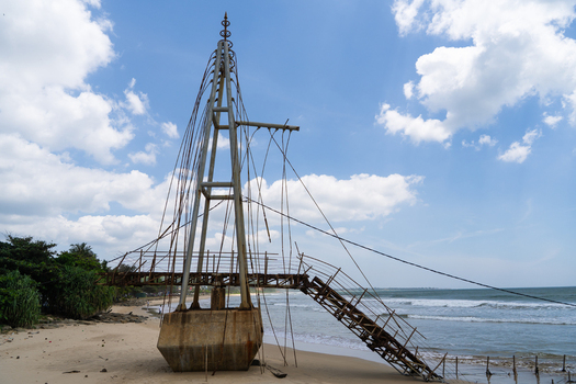 View of a weathered metal bridge structure on a sandy beach with ocean waves and tropical trees in Matara Southern Province Sri Lanka.