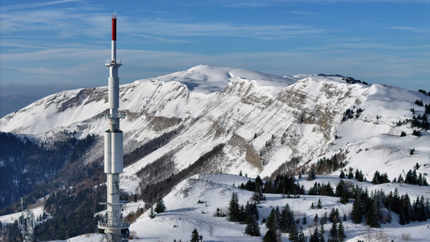 Aerial view of a towering communications tower piercing the crisp air against the backdrop of snow laden mountains their peaks dusted with sparkling white Gex Auvergne Rhône Alpes France. Aerial view of a towering communications tower piercing the crisp air against the backdrop of snow laden mountains their peaks dusted with sparkling white Gex Auvergne Rhône Alpes France.