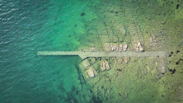 Aerial view of the rugged coastline meeting the turquoise sea where a stone pier stretches out a stark contrast against the clear waters Carantec Brittany France.