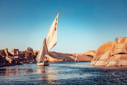 View of a traditional felucca sailboat with a large white sail navigating the Nile River past rocky banks and desert hills under a clear blue sky in Aswan Aswan Governorate Egypt. View of a traditional felucca sailboat with a large white sail navigating the Nile River past rocky banks and desert hills under a clear blue sky in Aswan Aswan Governorate Egypt.