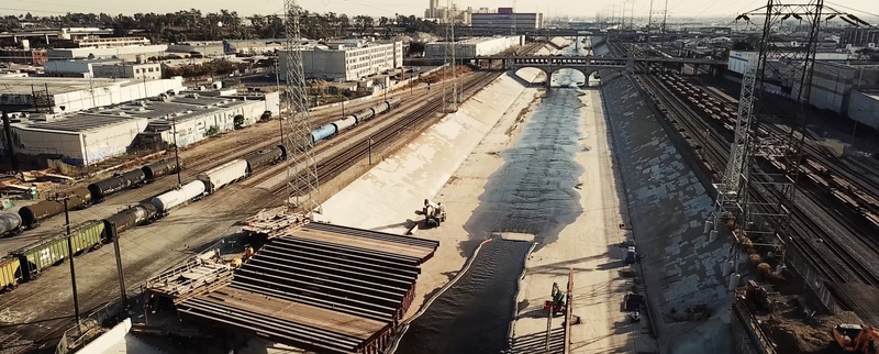 Aerial view of the concrete lined Los Angeles River channel with freight trains on adjacent tracks industrial buildings and a bridge in Los Angeles California United States.